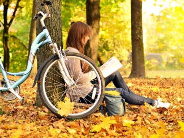 Eine junge Studentin sitzt im Park an einen Baum gelehnt und ist in ein Buch vertieft. Im Vordergrund steht ihr Fahrrad.
